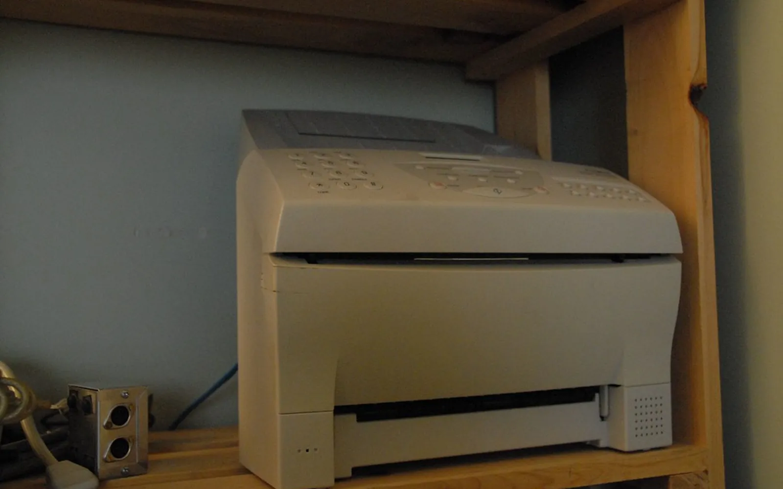 A desktop fax machine on a wooden office shelf with a phone line socket beside it