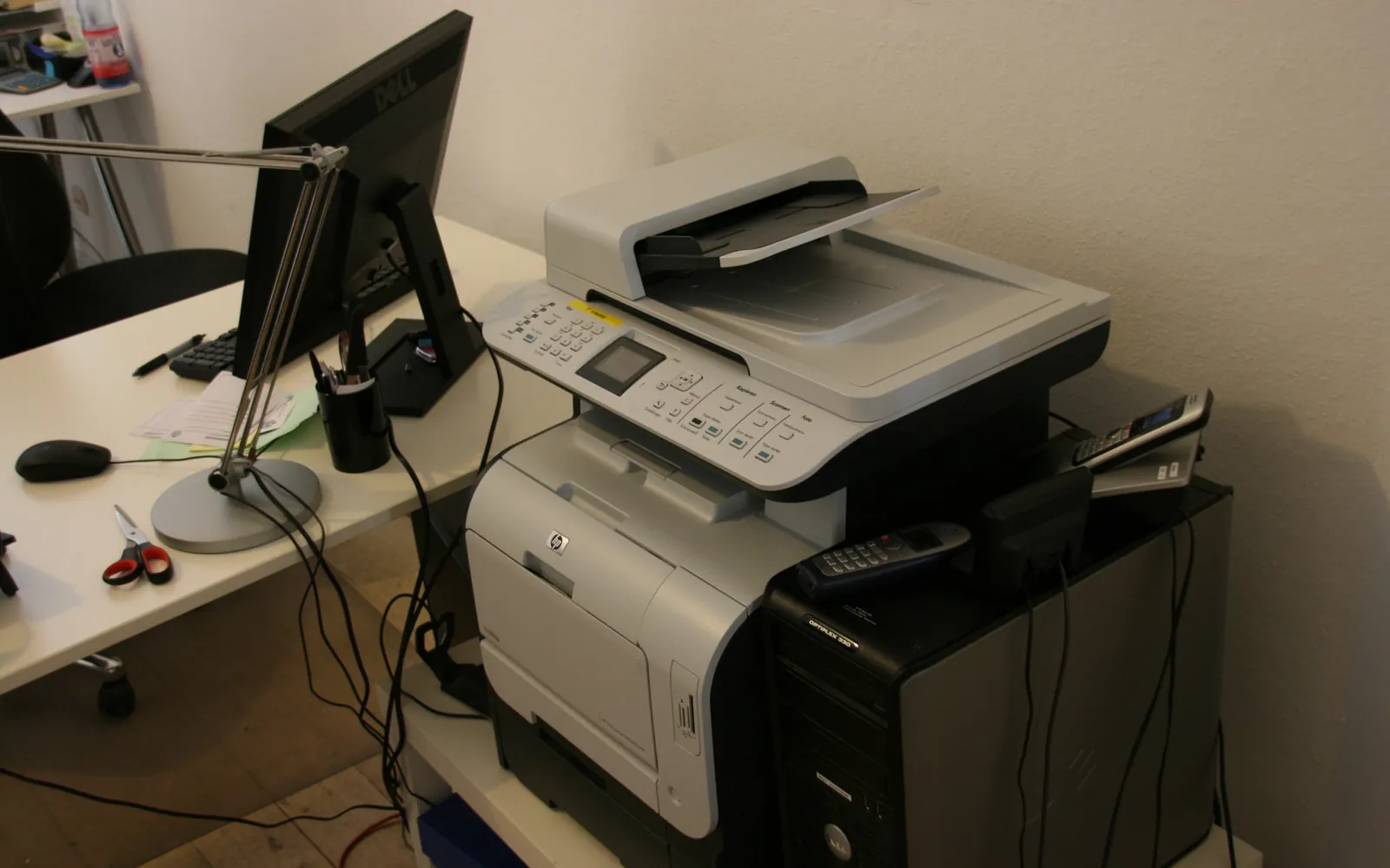 A multifunction office printer with document feeder next to a desk and monitor