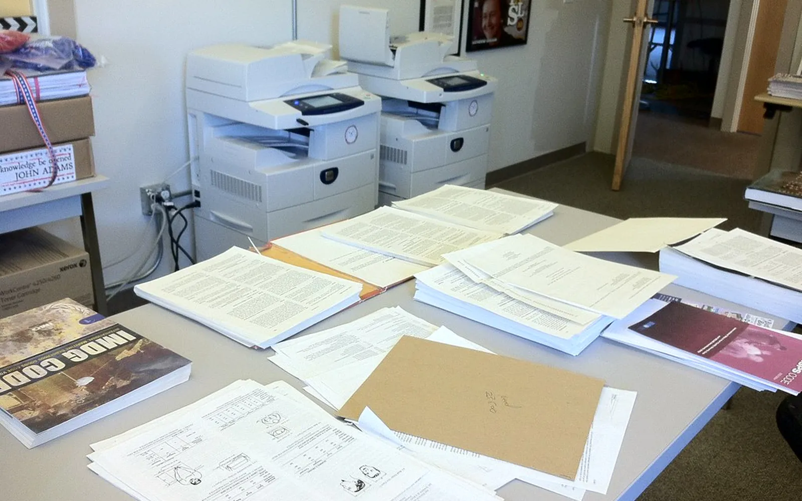 Two office multifunction printers behind a table covered with stacks of documents waiting to be scanned